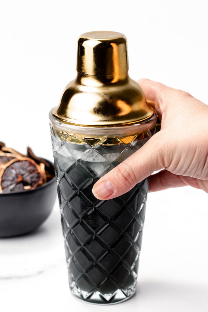 A hand holding a diamond-patterned cocktail shaker with a gold lid; a bowl with dried citrus slices and hints of apple cider punch sits in the background.