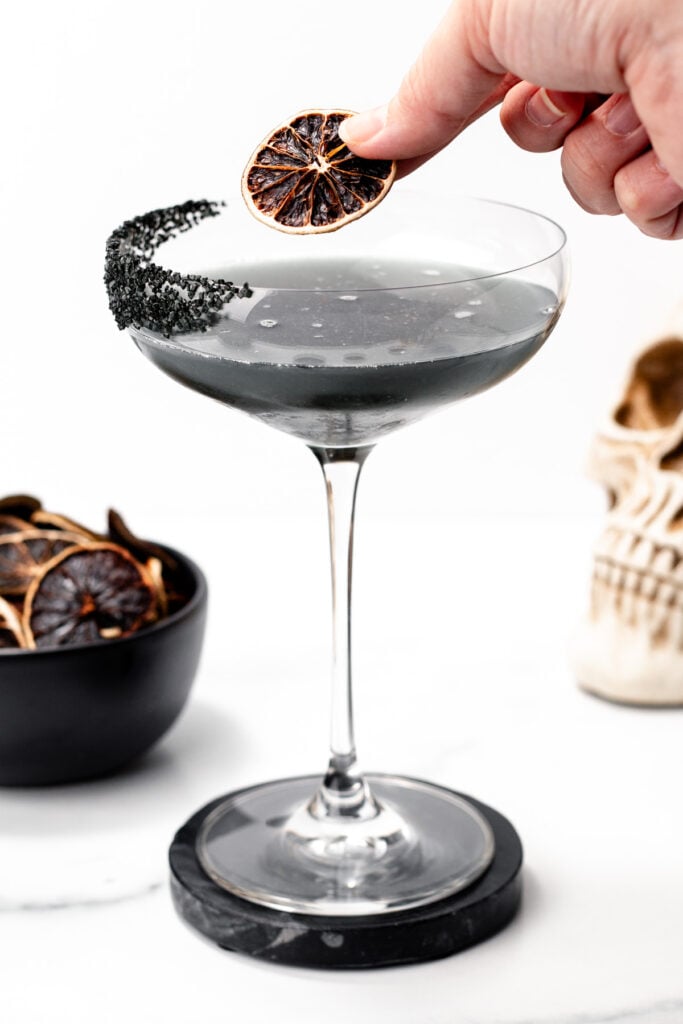 A hand places a dried citrus slice garnish on a black cocktail in a coupe glass with a black salt rim. A bowl of dried citrus, a decorative skull, and hints of apple cider punch inspiration are in the background.