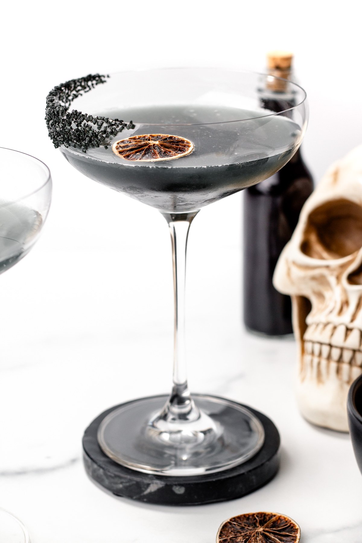 A coupe glass filled with a dark apple cider punch, garnished with a dried lime slice and black sugar on the rim; a skull decoration and bottle are in the background.