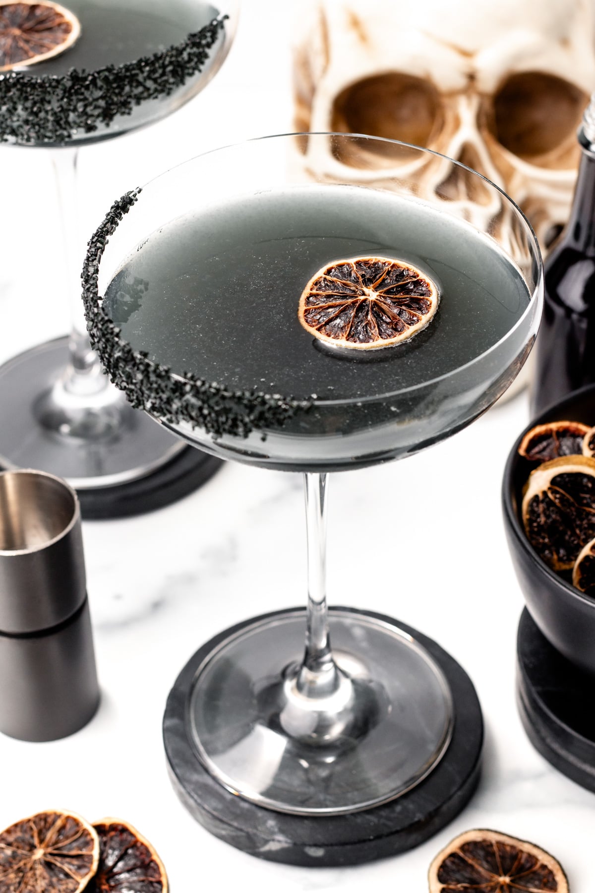 A cocktail in a coupe glass with a black sugar rim and dried citrus garnish, inspired by apple cider punch, sits on a black coaster with a skull decoration in the background.