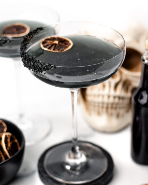 A coupe glass with a black cocktail, black sugar rim, and a dried citrus slice garnish evokes a mysterious twist on apple cider punch. Another glass, dried citrus slices, a dark bottle, and skull decor complete the moody scene.