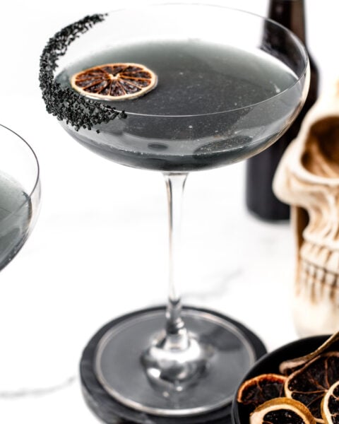 A coupe glass filled with a dark gray apple cider punch cocktail, garnished with a dried citrus slice and black salt on the rim. A skull decoration and more dried citrus slices are in the background.