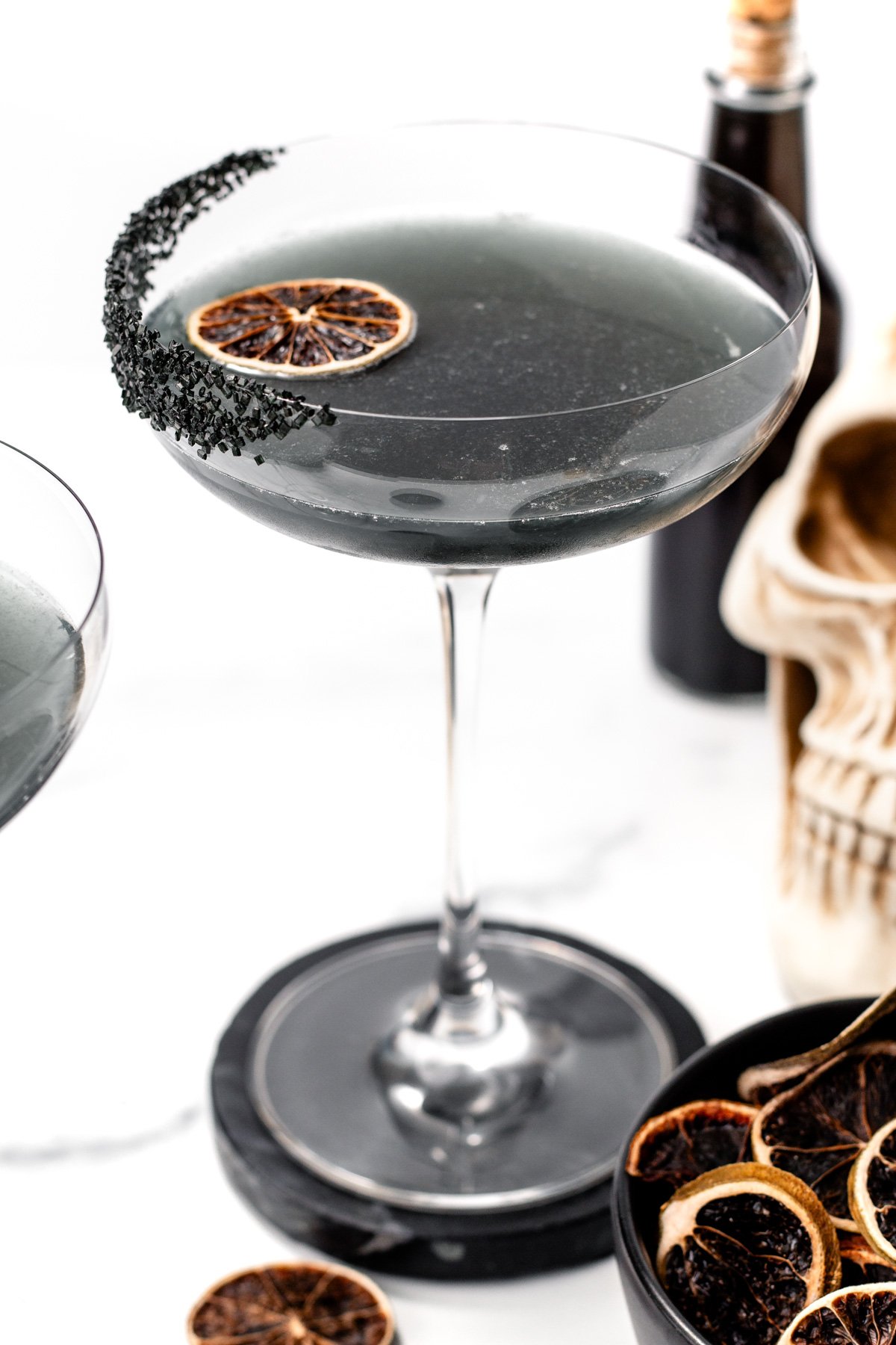 A coupe glass filled with a dark gray apple cider punch cocktail, garnished with a dried citrus slice and black salt on the rim. A skull decoration and more dried citrus slices are in the background.