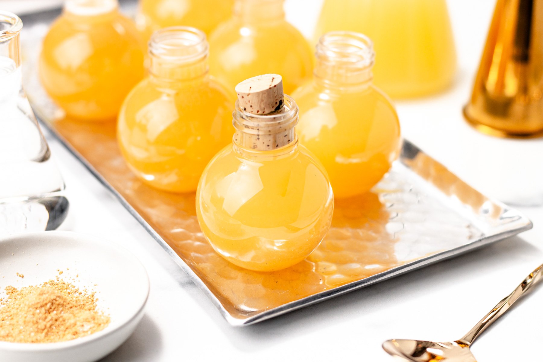 Round glass bottles filled with yellow apple cider punch, one with a cork stopper, are arranged on a rectangular metal tray with other bar tools nearby.