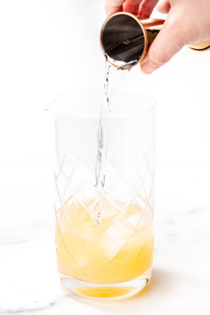 A hand pours a clear liquid from a jigger into a mixing glass containing a yellow apple cider punch, set on a white surface.