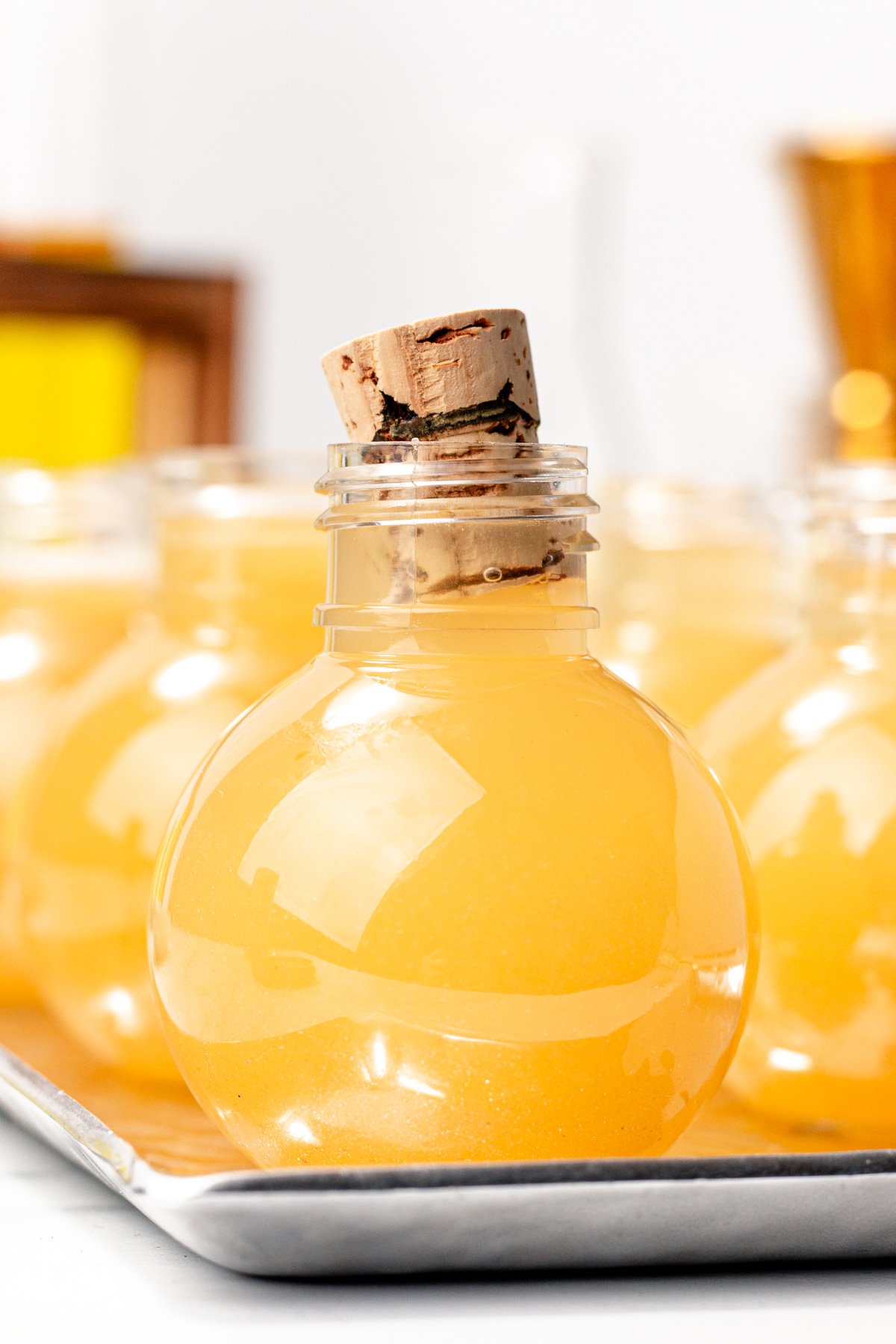 A round glass bottle filled with yellow-orange apple cider punch, sealed with a cork, sits on a metal tray with similar bottles in the background.