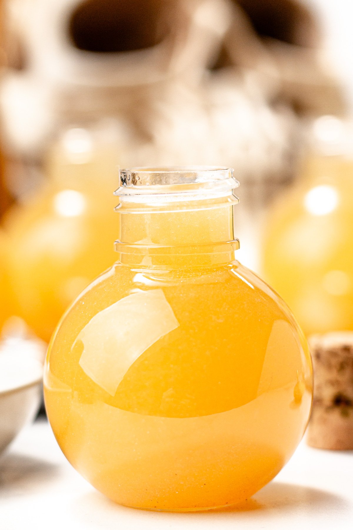A small round glass bottle filled with a thick, yellow-orange apple cider punch, shown with its cork stopper placed beside it.