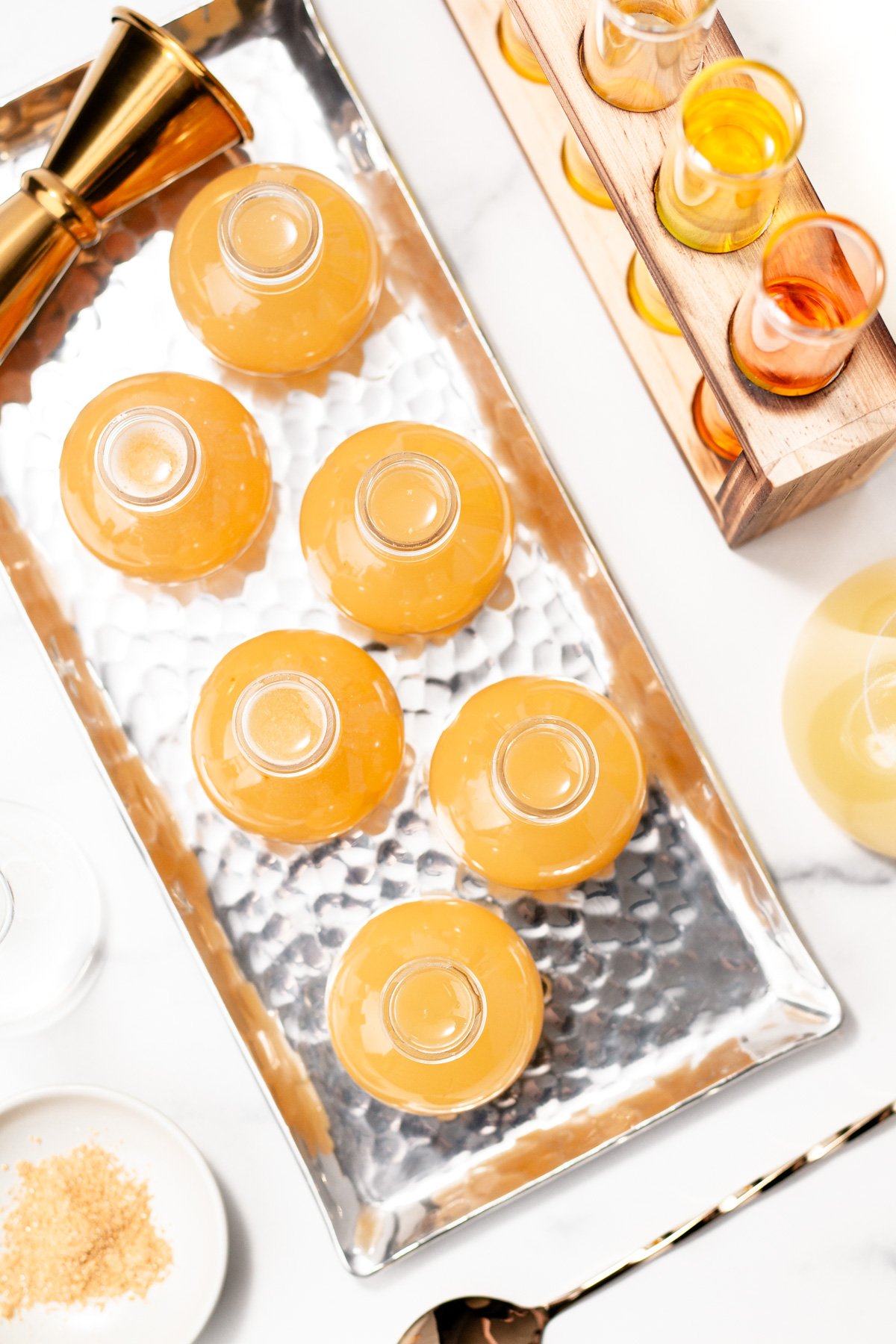 Six filled cocktail glasses, perfect for serving apple cider punch, are arranged upside down on a rectangular hammered metal tray, with a jigger and a wooden shot glass holder visible nearby.