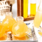 Three round glass bottles filled with yellow apple cider punch are displayed on a tray, with a cork stopper nearby and laboratory glassware and a skull in the background.