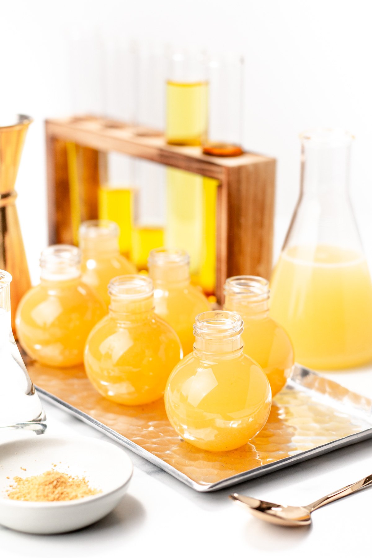 Clear round bottles filled with orange apple cider punch are arranged on a tray, with laboratory glassware containing similar liquid in the background.