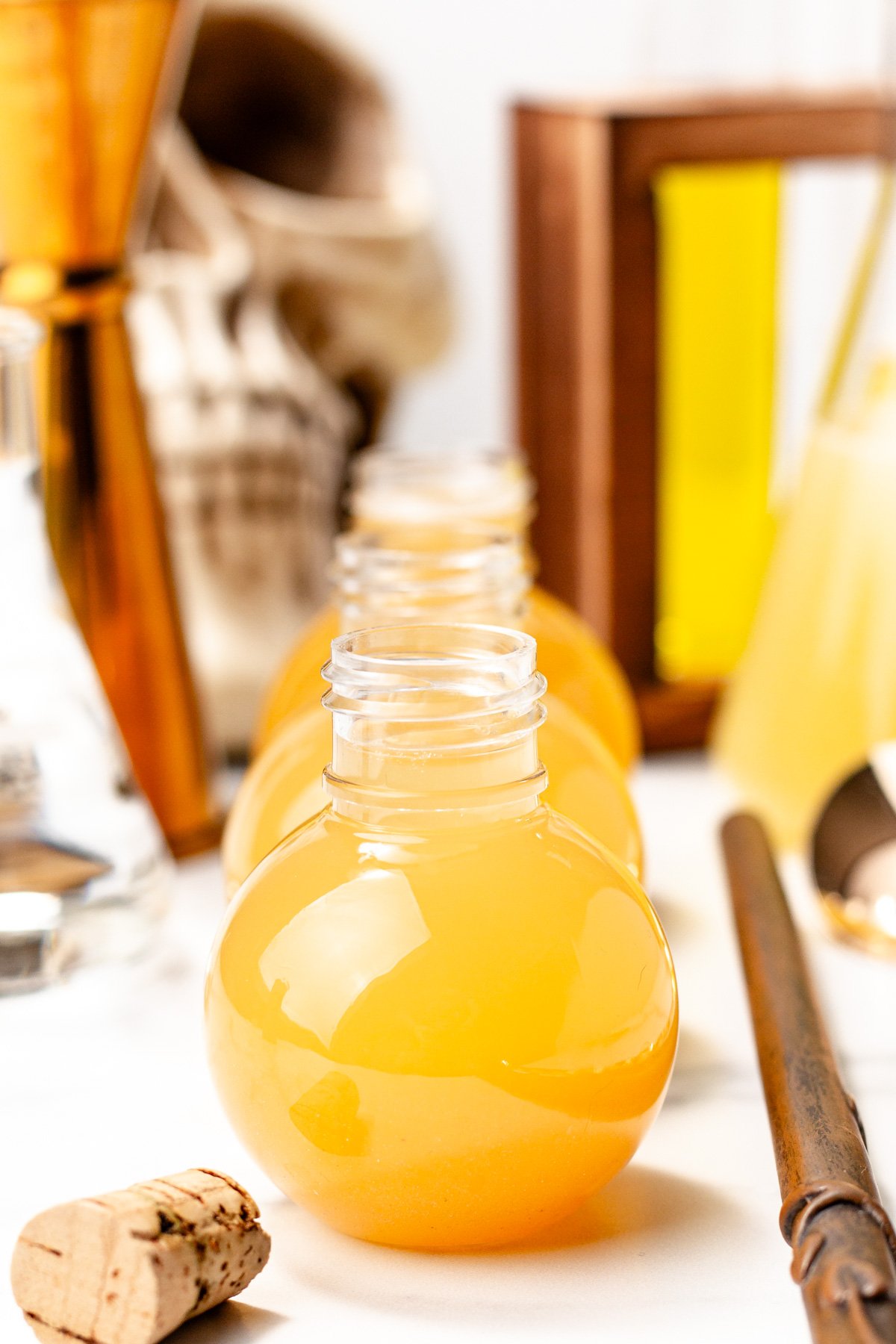Round glass bottles filled with orange liquid sit on a table with cocktail tools and a cork nearby; a skull decoration is visible in the background, hinting at apple cider punch ready for any festive gathering.