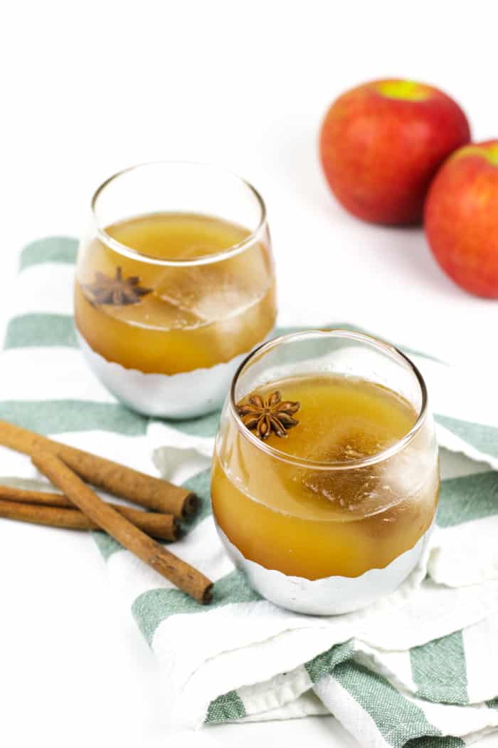 Two glasses of spiced apple cider rum punch with ice and star anise, placed on a striped cloth with cinnamon sticks and apples in the background.