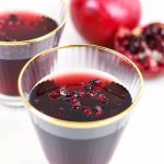 Two glasses of dark red pomegranate jelly with pomegranate seeds on top evoke the rich hue of a Port Wine Cocktail; a whole and a cut pomegranate are in the background.