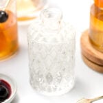 A textured glass bottle with clear liquid sits on a white surface, surrounded by two glasses of amber liquid with cherries and a small dish of dark cherries in syrup—perfect for showcasing homemade simple syrup creations.