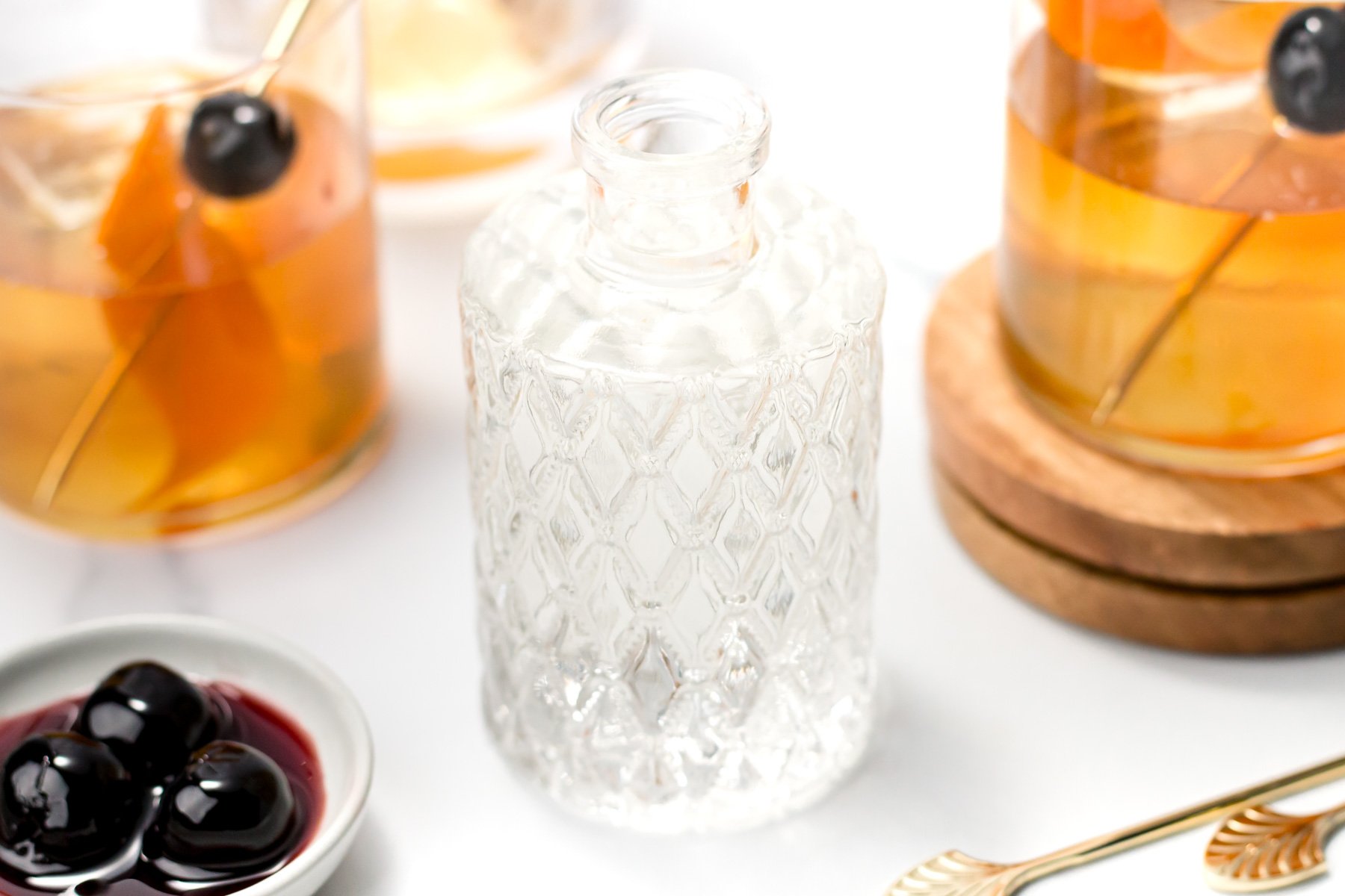 A textured glass bottle with clear liquid sits on a white surface, surrounded by two glasses of amber liquid with cherries and a small dish of dark cherries in syrup—perfect for showcasing homemade simple syrup creations.