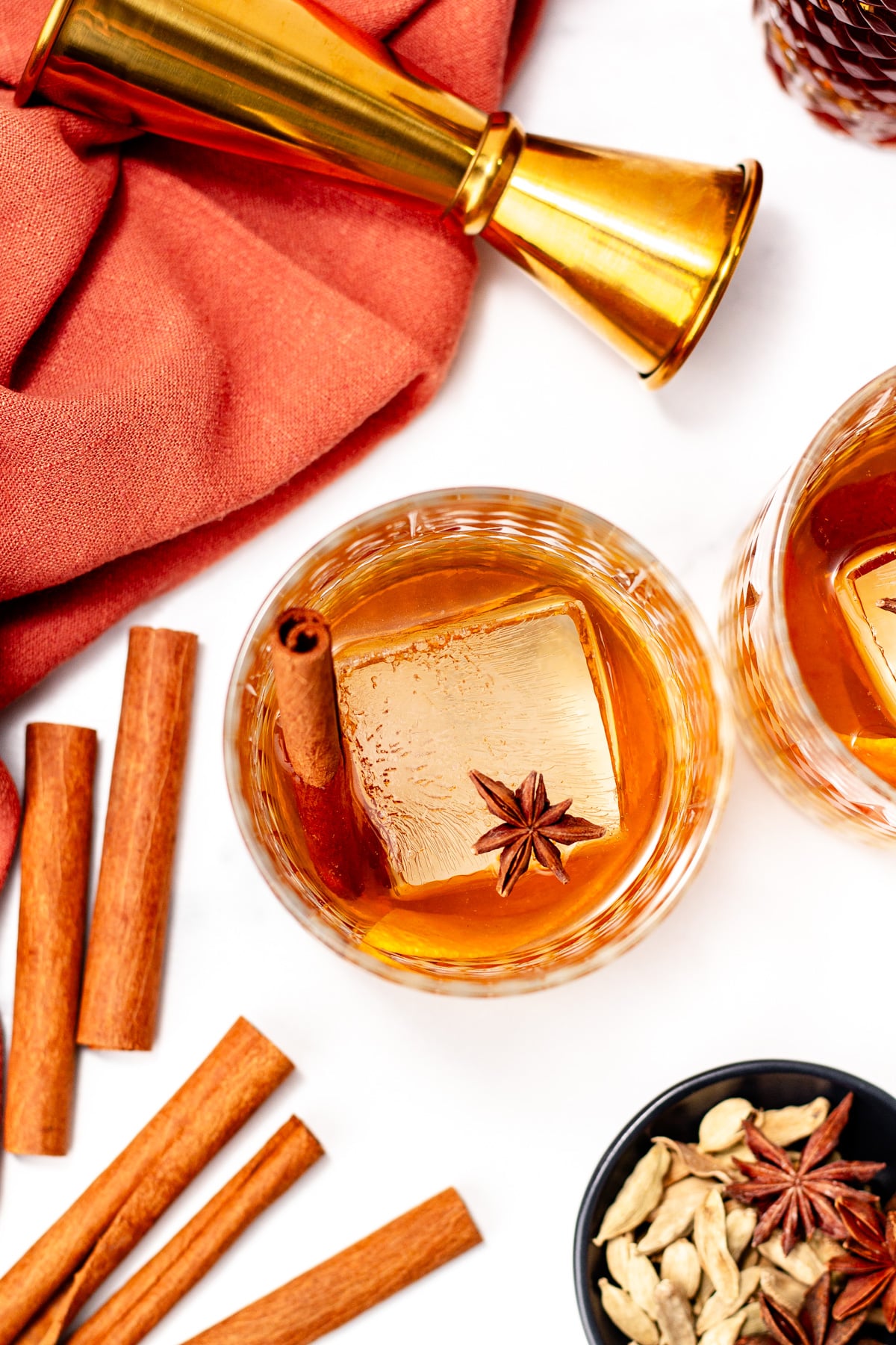 Two glasses of chai cocktail with spiced whiskey and large ice cubes, garnished with cinnamon sticks and star anise. A jigger, loose spices, and an orange cloth are nearby.
