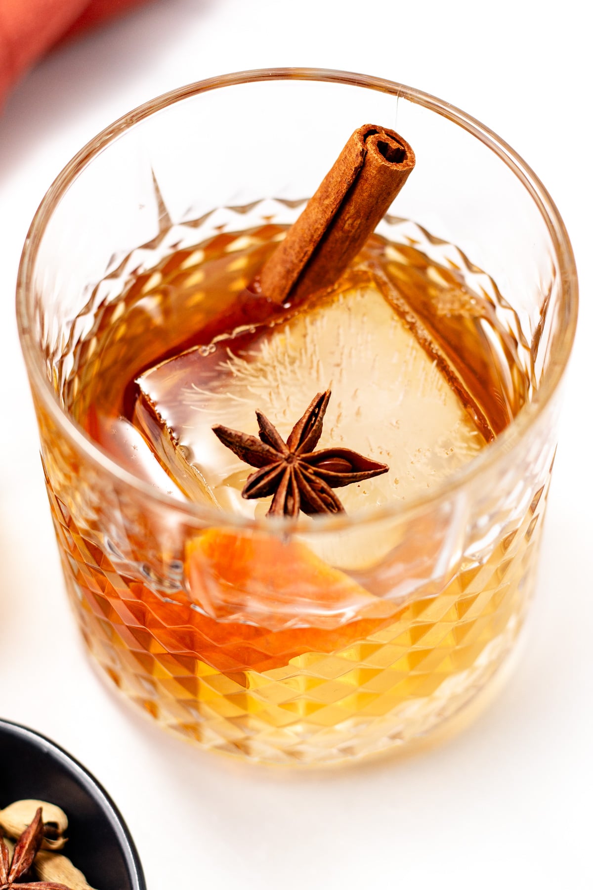 A glass of amber-colored chai cocktail with a large ice cube, garnished with a cinnamon stick and a star anise.