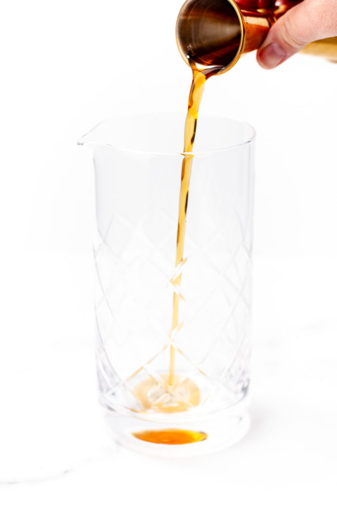 A hand pours a rich brown liquid, blending a chai cocktail from a small metal cup into a large, empty, clear glass mixing vessel against a white background.