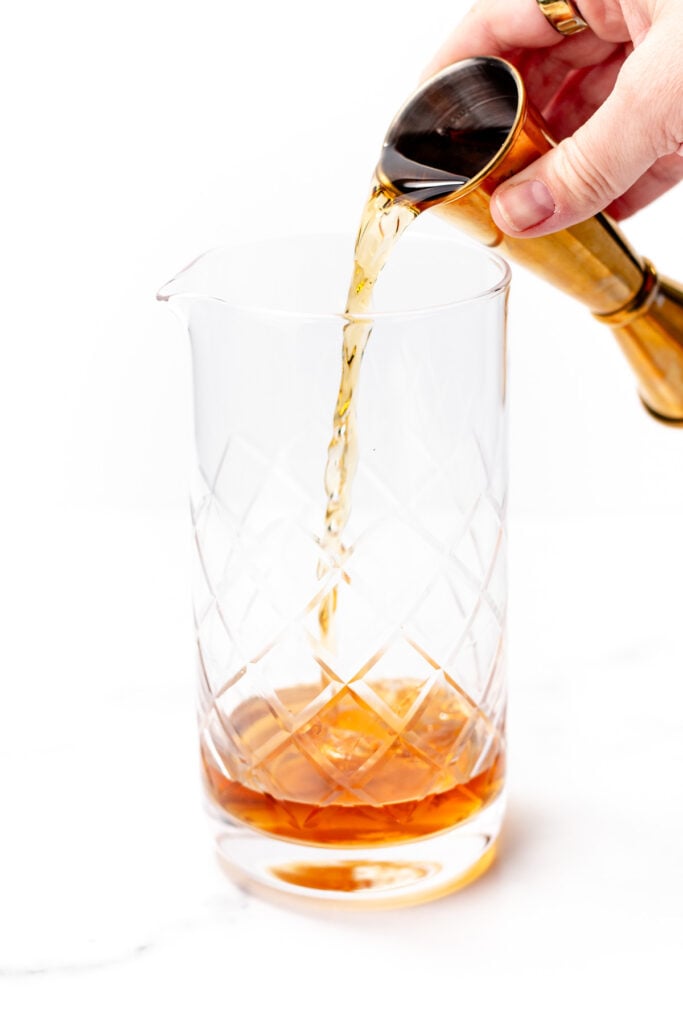 A hand pours amber liquid from a jigger into a clear, patterned glass mixing pitcher on a white background, crafting a chai cocktail.