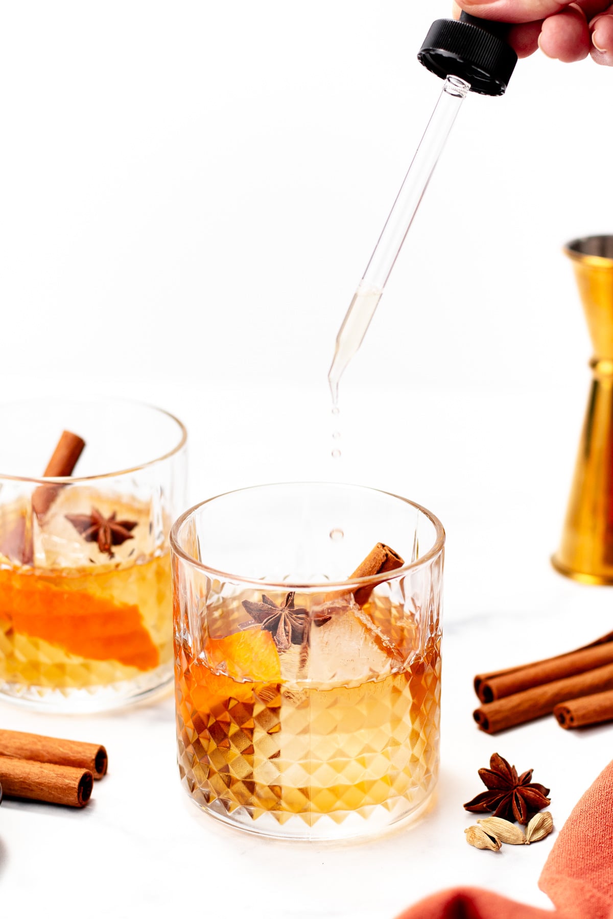 A person adds liquid with a dropper to a chai cocktail in a glass garnished with orange peel, star anise, and a cinnamon stick. Another similar glass and cocktail tools are nearby.