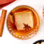 A glass of amber-colored chai cocktail with a large ice cube, garnished with a cinnamon stick and star anise, sits on a white surface next to a red cloth.