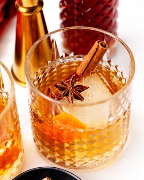 A glass of chai cocktail garnished with a cinnamon stick, star anise, and orange peel, with a patterned bottle and jigger in the background.