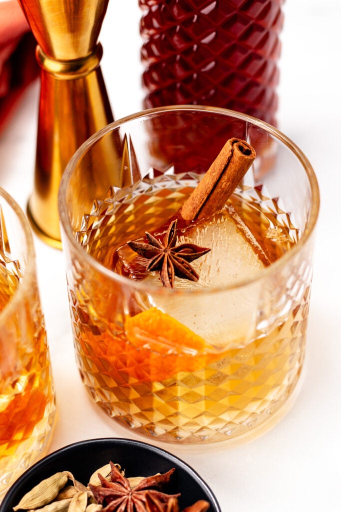 A glass of chai cocktail garnished with a cinnamon stick, star anise, and orange peel, with a patterned bottle and jigger in the background.