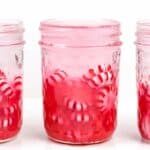 A glass jar partially filled with red and white peppermint candies for homemade peppermint vodka sits between two similar jars against a white background.
