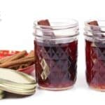Two glass jars filled with a dark liquid, each with a cinnamon stick inside, sit next to a jar and a bowl containing more cinnamon sticks—perfect for crafting homemade cinnamon whiskey—and two metal lids.