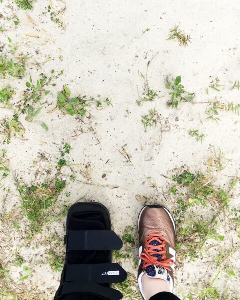A person, grounded by a medical boot on the left foot and a sneaker on the right, stands on a sandy ground with patches of grass, contemplating the challenges of flying with an injury.