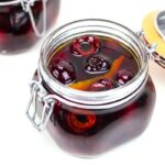 A glass jar filled with dark liquid, homemade cocktail cherries, and an orange peel, with the lid open; another similar jar is partially visible in the background.