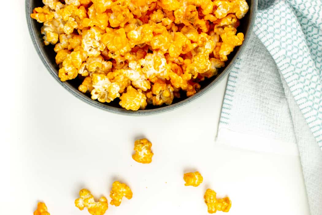 a bowl of cheddar cheese popcorn with kernels spilling out of the side of the bowl onto a white surface