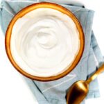 a top view of a gold spoon and a gold bowl containing the best homemade whipped cream on a blue tea towel