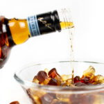 whiskey being poured into a clear glass bowl of cola gummy candy on a white background