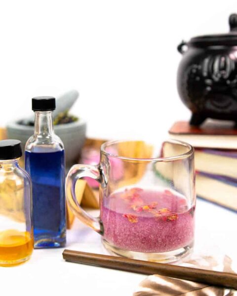 the amortentia potion cocktail from harry potter surrounded by potion-sized bottles of colorful cocktail ingredients, a magic wand, a feather, a stack of books and a small cauldron