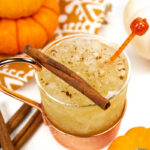 a pumpkin spice mule in a glass and copper mug with a cinnamon stick on top, surrounded by mini pie pumpkins