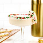 a candy cane cookie garnishing a sugar cookie martini next to baked and frosted sugar cookies and a cocktail shaker