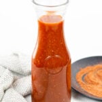 a carafe of homemade Bloody Mary mix with a kitchen towel and a plate of Bloody Mary rim salt