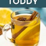 A glass mug of bourbon hot toddy with a lemon slice, cinnamon stick, and star anise, set beside a lemon wedge on a white surface. Text reads "Hot Toddy" and "Get the Recipe.
