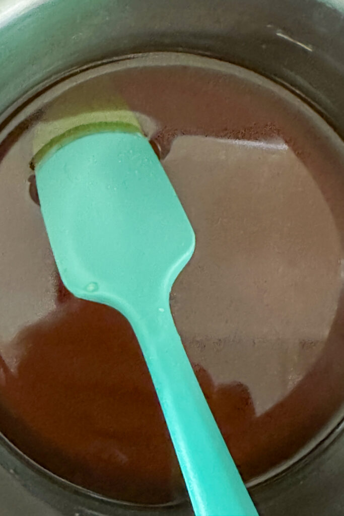 A teal spatula rests in a metal bowl containing melted chocolate, ready to be stirred with a drizzle of brown sugar simple syrup.