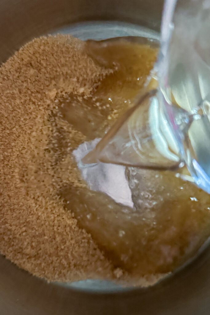A close-up of granulated brown sugar in a bowl with water being poured over it from a glass container, illustrating the first step in making demerara syrup.