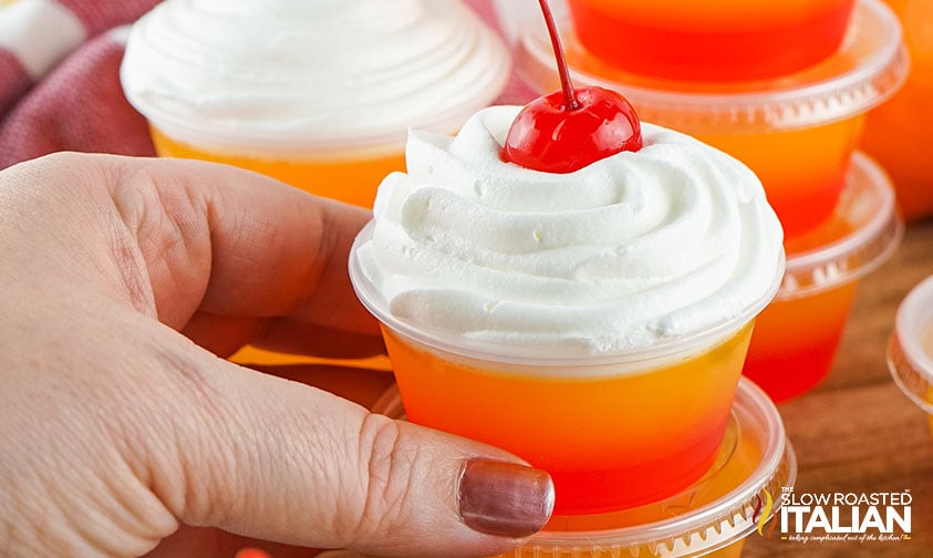 A hand holds a small plastic cup filled with orange and yellow layered gelatin, topped with whipped cream and a cherry—perfect inspiration for creative jello shot recipes. Other similar cups are visible in the background.