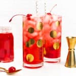 two dirty Shirley Temple cocktails with a jigger, a spoon and a jar of maraschino cherries.