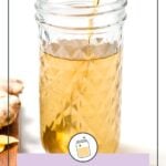 A glass jar filled with light brown ginger syrup sits on a white surface, a spoon inside and pieces of ginger nearby. Text reads “Ginger Simple Syrup.”.