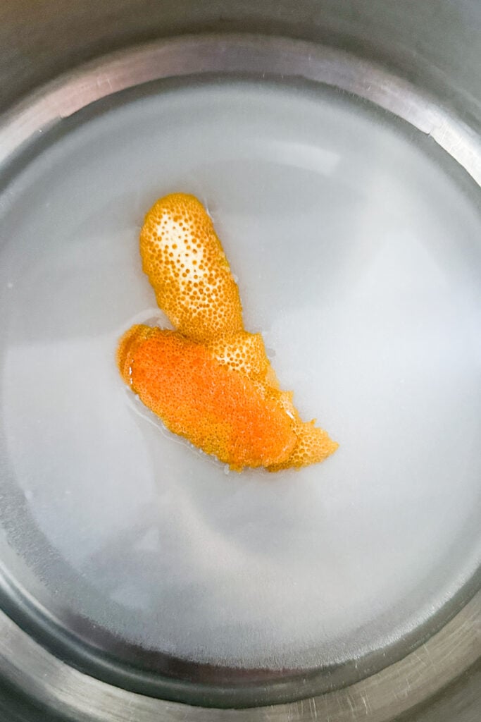 A close-up of a metal pot containing water with two pieces of orange turmeric root floating on the surface, perfect for infusing into an old-fashioned simple syrup.