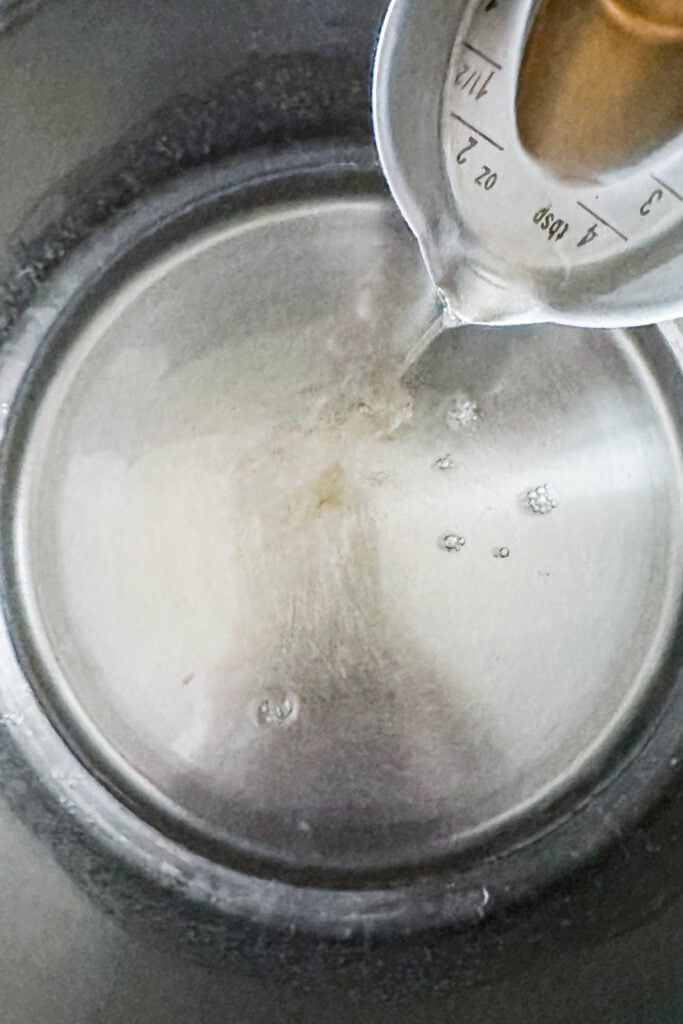 A measuring cup pours water and old-fashioned simple syrup into a stainless steel bowl, with droplets and reflections visible on the metal surface.