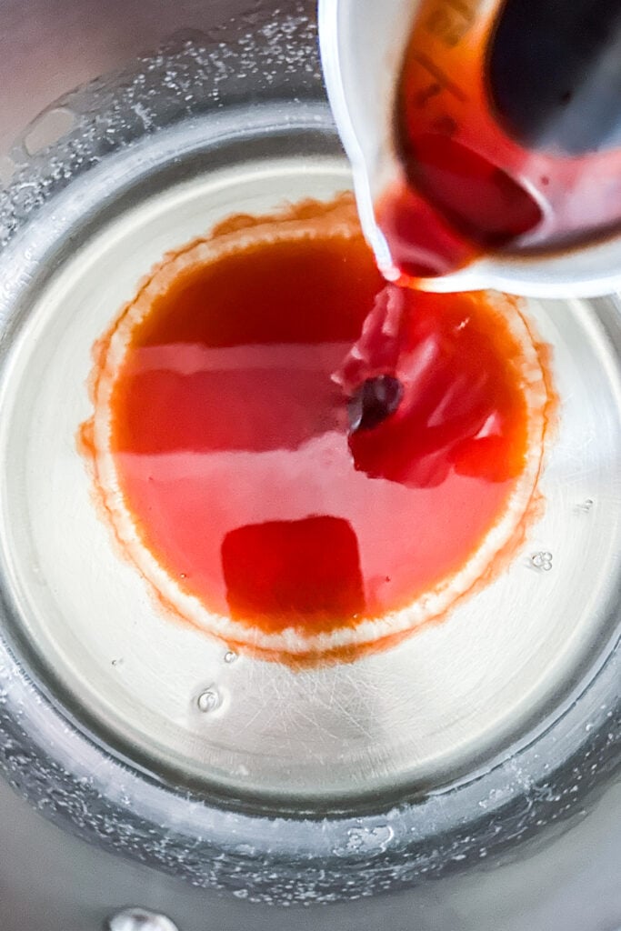 Old-fashioned simple syrup is being poured from a measuring cup into a metal bowl, forming a vibrant red ring on the surface.