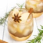 Rosemary sprigs, cinnamon sticks and star anise lay on a white wooden background alongside two stemless wineglasses filled with ice and golden liquid garnished with apple slices and spices