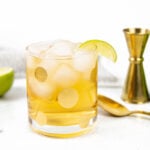 A gray and white dish towel wraps around a half of a lime, a gold spotted glass, a gold spoon and cocktail jigger and a Seagram's Ginger Ale