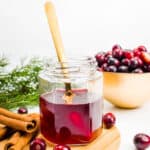 A gold spoon rests in a hexagon jar of cranberry-colored simple syrup with whole cranberries, cinnamon sticks, evergreen boughs and a golden bowl of cranberries around it